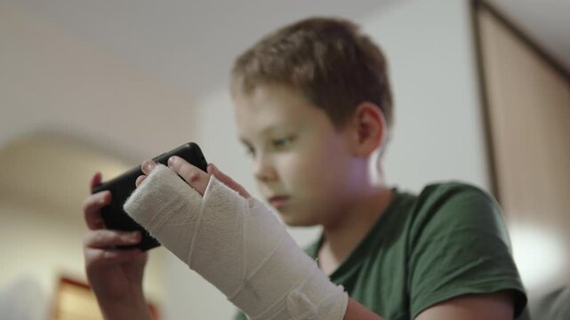 Boy playing smartphone during wearing cast on hand. Home routine and pastime after arm fracture during fixation period. Focused portrait of child during remote learning after injury.