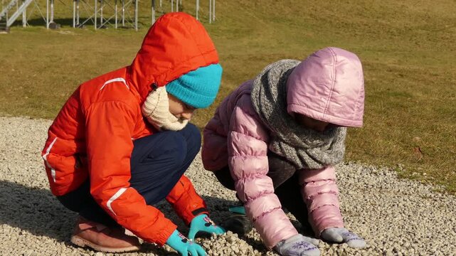 Two little beautiful girls walk in winter clothes collect gravel on the path.