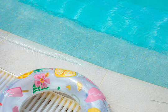 Colorful inflatable swim ring with flamingo and fruit print at pool edge, turquoise pool water and stone coping visible above