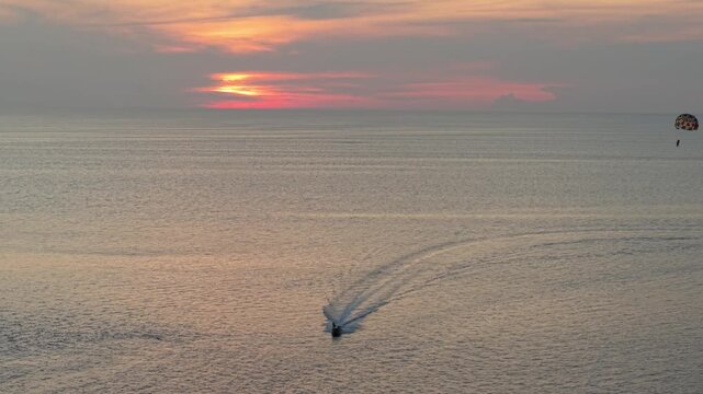 A boat moves swiftly across the calm water while a parachute glider rises into the air against a vibrant sunset backdrop, creating a dynamic scene of adventure and leisure