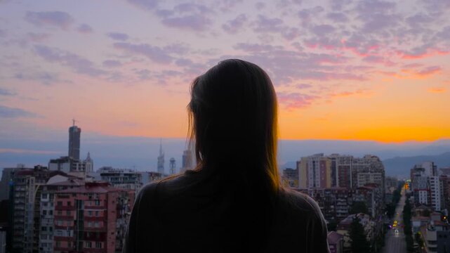 Back view: woman silhouette is standing on the balcony and looking around against the sunset, sunrise sky over the city. Lonely, urban, dramatic and freedom concept