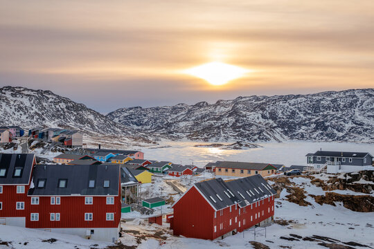 Arctic sunset landscape with Greenlandic residential houses, snow-covered hills, frozen sea water and mountain backdrop, Qaqortoq, Kujalleq, Greenland