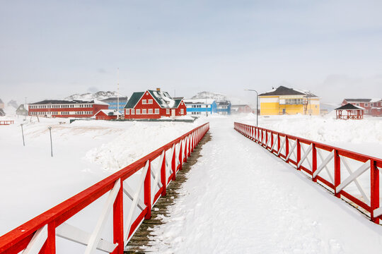 Snow-covered bridge and village street in Paamiut with red railing, colorful houses, overcast sky and arctic winter weather, Paamiut, Sermersooq, Greenland