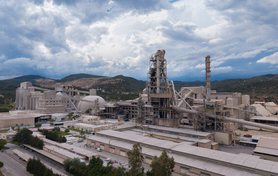 Aerial view of a large cement manufacturing plant featuring preheater tower, clinker storage dome, silos and industrial facilities surrounded by mountain landscape under dramatic cloudy sky