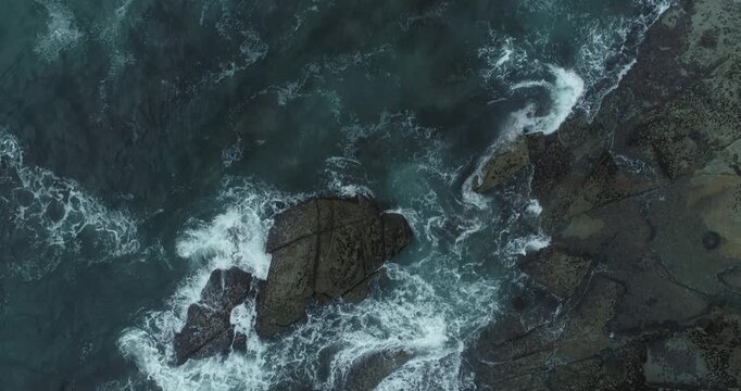 Dramatic drone photography of dark stones and white sea foam swirling in a turbulent ocean