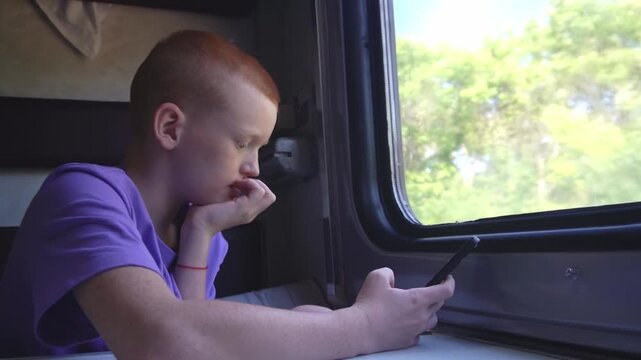 A red-haired teenager holds a phone in his hand and texts while sitting in his compartment during a train journey. High quality FullHD footage