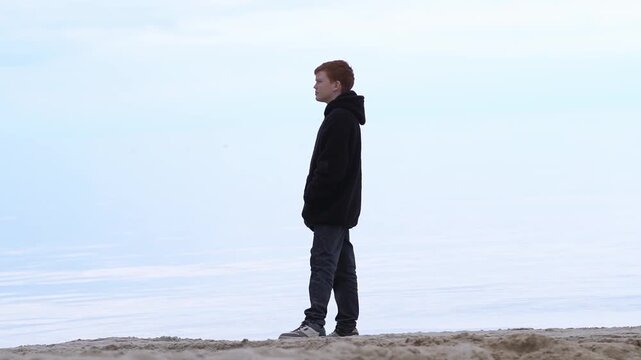 A teenage boy in a jacket and jeans strolls along the shoreline of a sea or ocean, the water and sky form a blurred horizon, creating an endless blue water that turns into clouds.