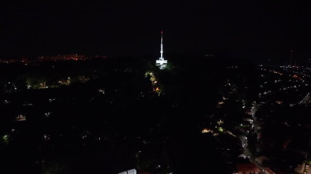 Night drone footage over Miskolc, Hungary, featuring the iconic Avas TV Tower rising above the city skyline. The video captures the illuminated tower, glowing streets, hillside neighborhoods