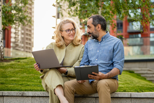 Middle-aged couple discussing work on laptop and tablet during a casual outdoor meeting in park