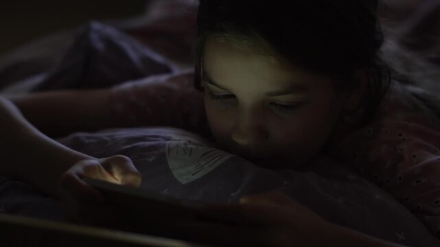 A teenage girl lies in bed before sleep and holds her phone in her hands scrolling through her online feed, which illuminates her face. High quality FullHD footage