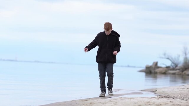 A teenage boy in a jacket and jeans walks along the coastline of the sea or ocean. High quality FullHD footage