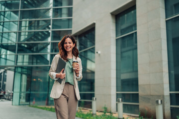 Fototapeta premium Professional businesswoman smiling, walking confidently with laptop and coffee, ready for work in an urban office area