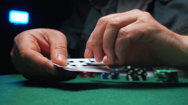 A male gambler with visible hands holding a deck of playing cards shows a two pairs combination while sitting at a green felt poker table surrounded by stacks of poker chips and dim lighting