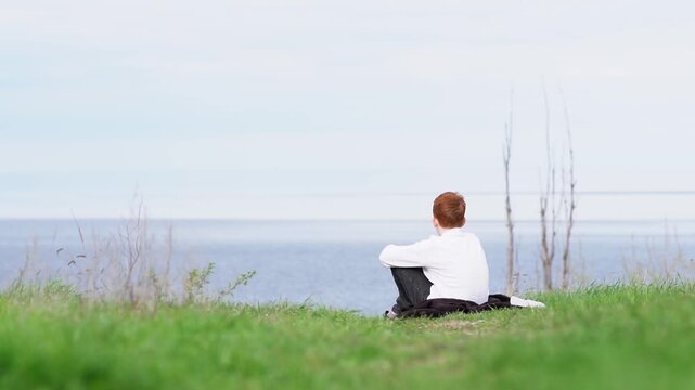 A guy is sitting on a hill overlooking a calm, serene ocean with a cloudless sky. High quality FullHD footage