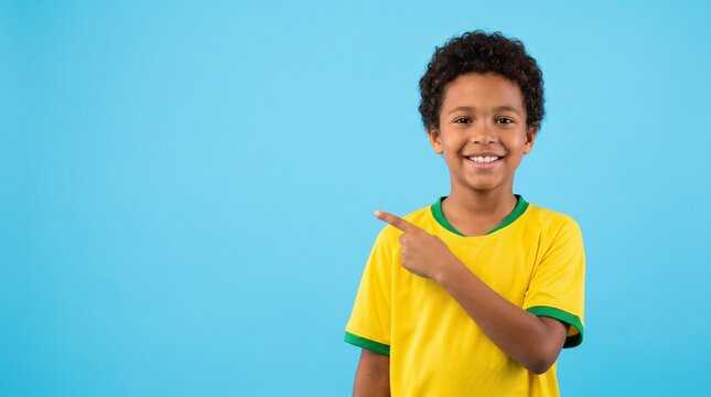 A cheerful young boy with curly hair wearing a yellow and green t-shirt smiles and points his finger toward copy space against a solid blue background, ideal for advertising or sports themes
