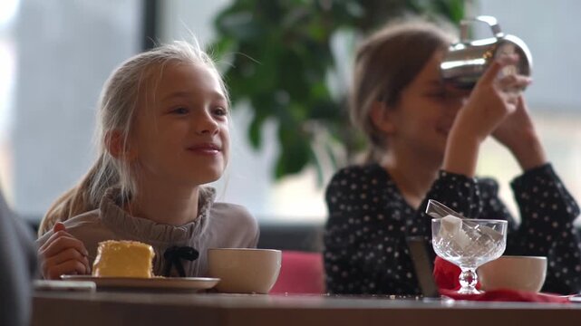 Little girls drink tea with dessert in a cafe, talk and indulge. A girl with a teapot in her hands drinks its spout. High quality FullHD footage