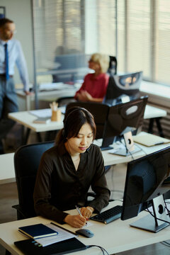 Vertical shot of Asian young adult woman working in call center office, wearing headset and taking notes at computer. Useful for customer support, team communication, service business