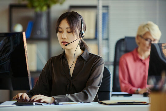 Medium shot of Asian young adult woman wearing headset, typing at computer in call center office, handling customer support and teamwork. Ideal for service business and CRM marketing