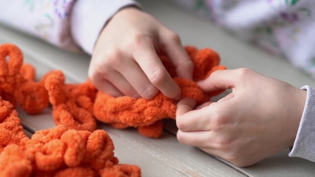 Little baby's fingers are knitted using orange plush yarn.. Children's hobby. The girl sits holding a woolen thread in her hands.
