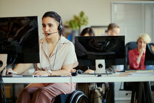 Young adult woman with disability working in call center, wearing headset and using computer in team office. Customer support and inclusive workplace concept for service business marketing