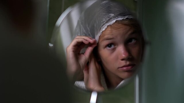 A girl looks at herself in a round mirror in a hotel bathroom and adjusts the clear shower cap on her hair. The teenager is performing her morning or evening body care routine.