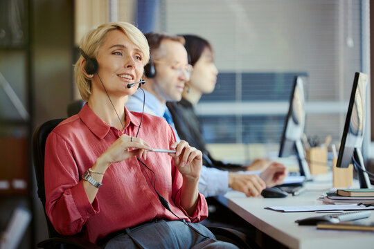 Medium shot of Caucasian young adult woman wearing headset, assisting customers in call center office with team support. Ideal for customer service, CRM, outsourcing marketing