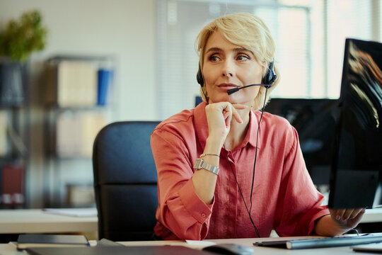 Middle aged Caucasian woman wearing headset, assisting customers and monitoring computer in call center office. Ideal for customer support, CRM, contact service marketing