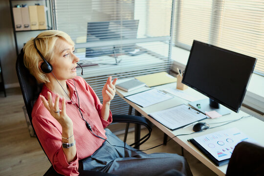 Young adult Caucasian female operator wearing headset, speaking with client and gesturing at desk in call center office. Ideal for customer support, CRM, service team marketing