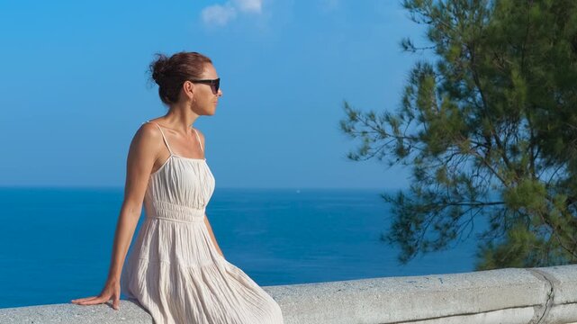 Woman sitting on wall enjoying ocean view. Relaxed female traveler wearing white dress and sunglasses sitting on coastal stone wall, gazing across serene blue seascape with pine tree