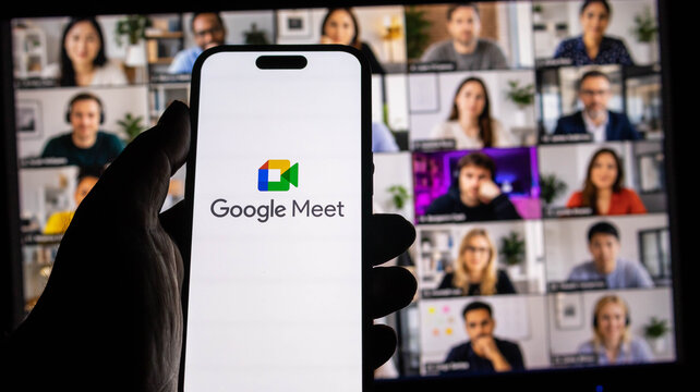 Closeup of a smartphone displaying the google meet app logo held in front of a computer screen showing a video conference