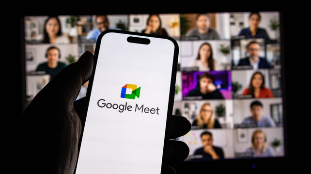 Closeup of a hand holding a smartphone displaying the google meet app logo with a video conference in the background