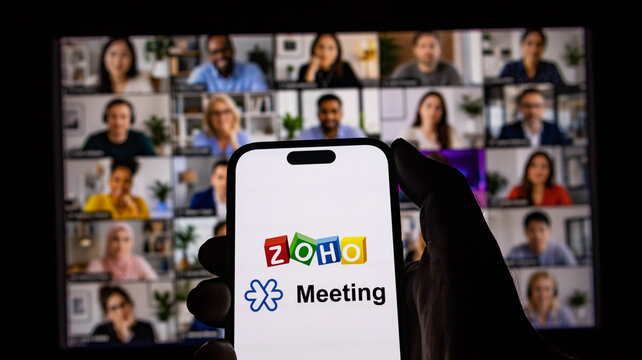 Closeup of a hand holding a smartphone displaying the zoho meeting app logo in front of a video conference screen