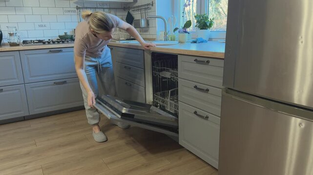 Female opening dishwasher door and sliding out lower rack to check dishes inside in modern kitchen.