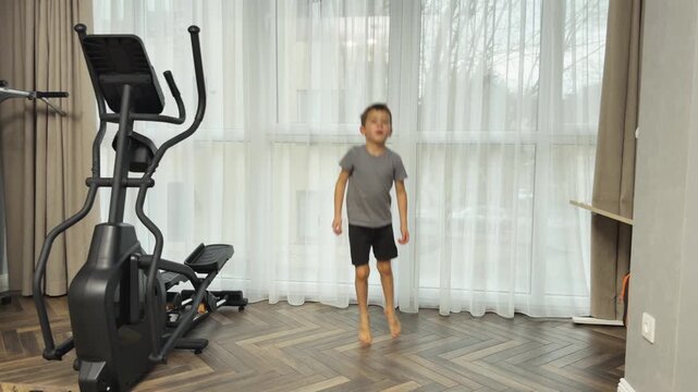 Small boy in shorts and t shirt performs two foot jumps forward and backward in living room near window and elliptical trainer. Home activity, playful fitness and energy release for children.