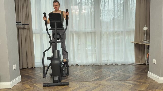 Woman performs forward stepping on elliptical machine in living room, working with arms and legs together. Home cardio exercise for endurance, coordination and overall fitness.