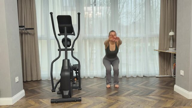 Female exercising in living room doing squats near cardio trainer equipment.