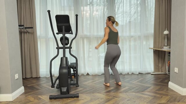 Woman performs dynamic jumps with 180 degree turns in bright living room near elliptical trainer. Home cardio routine improving coordination, balance and agility.