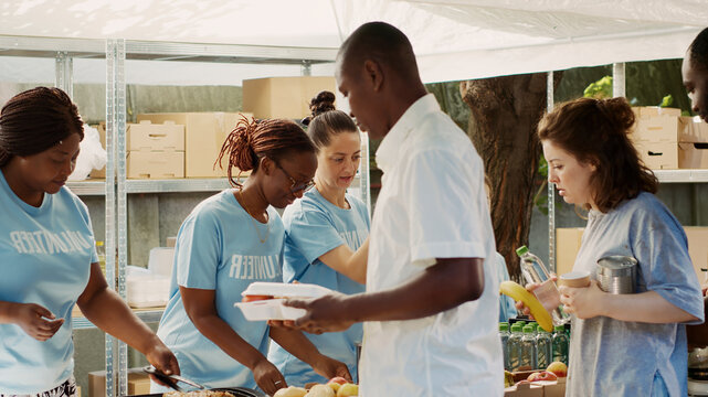 Multiracial volunteer ladies serve meals and non-perishable products to hungry and homeless people, friendly charity workers handing out free food to the poor and needy.