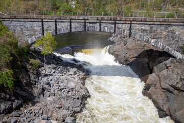 Obraz premium Old stone dam and arched bridge channel turbulent river rapids into a cascading waterfall through a rocky gorge, surrounded by spring foliage and rugged cliffs in bright daylight in N Walpole, NH