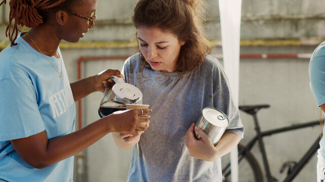 African American female volunteer serving a cup of coffee to homeless caucasian lady, friendly black woman at outdoor food bank sharing nourishments to the needy and less fortunate.