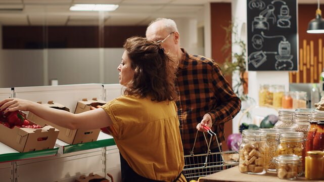 Zero waste supermarket vendor assisting elderly client with his shopping, adding needed food items to shopping basket, storekeeper helping senior customer in local neighborhood shop.