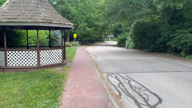 First person tracking shot moving past a wooden gazebo on a quiet suburban street