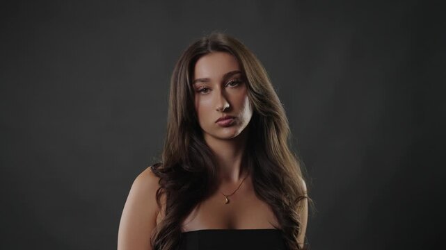 Confident young woman with long dark hair posing on a solid gray background. Her natural beauty and self assurance are evident in her calm expression and direct gaze. This studio portrait highlights h