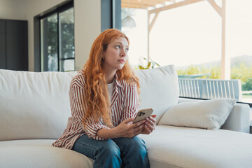 Fototapeta premium Redhead woman sits on a white couch in a modern living room, looking contemplatively out a large window while holding her smartphone, reflecting on thoughts or awaiting something