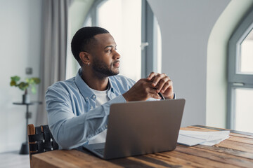Fototapeta premium Young black man at a wooden desk, thoughtfully gazing away from his laptop while working remotely in a modern apartment home office, planning and concentrating on a project
