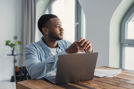 Young black man at a wooden desk, thoughtfully gazing away from his laptop while working remotely in a modern apartment home office, planning and concentrating on a project