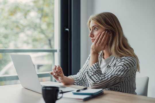 Young woman procrastinating during remote work, looking out the window, showing lack of motivation and distraction from her laptop workstation at a desk