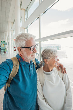 Senior couple embracing and happily smiling, looking out a large window at an airport terminal, anticipating an upcoming journey or retirement vacation