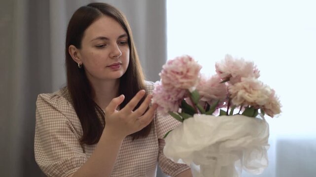 Serene young woman arranges pink peonies in a vase wearing gingham dress. Soft sunlight streams through window in bright home interior setting.
