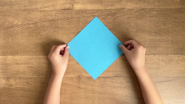 Child Hand Placing Light Blue Origami Paper on Wooden Table, 4K